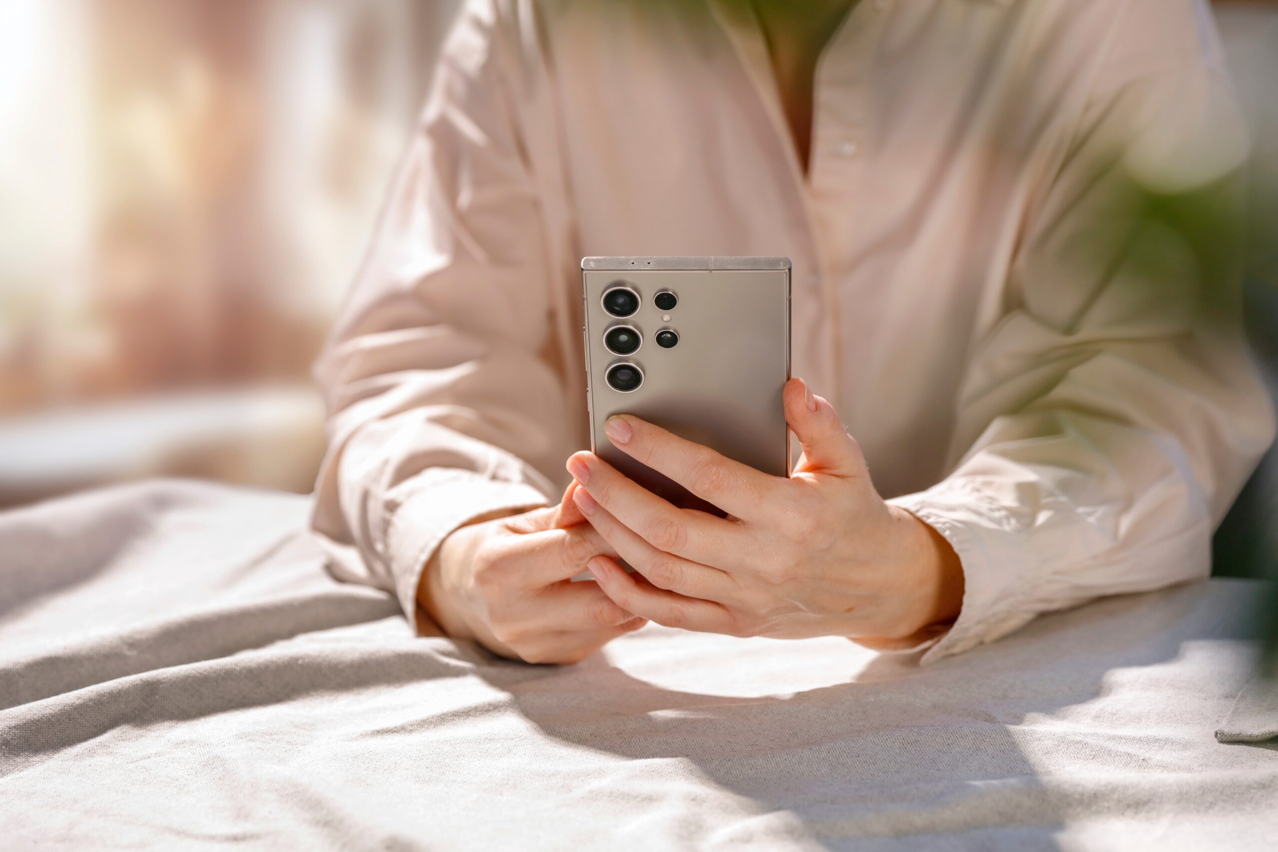 `alt="A person operating a smartphone in a space where sunlight filters through the trees. "`