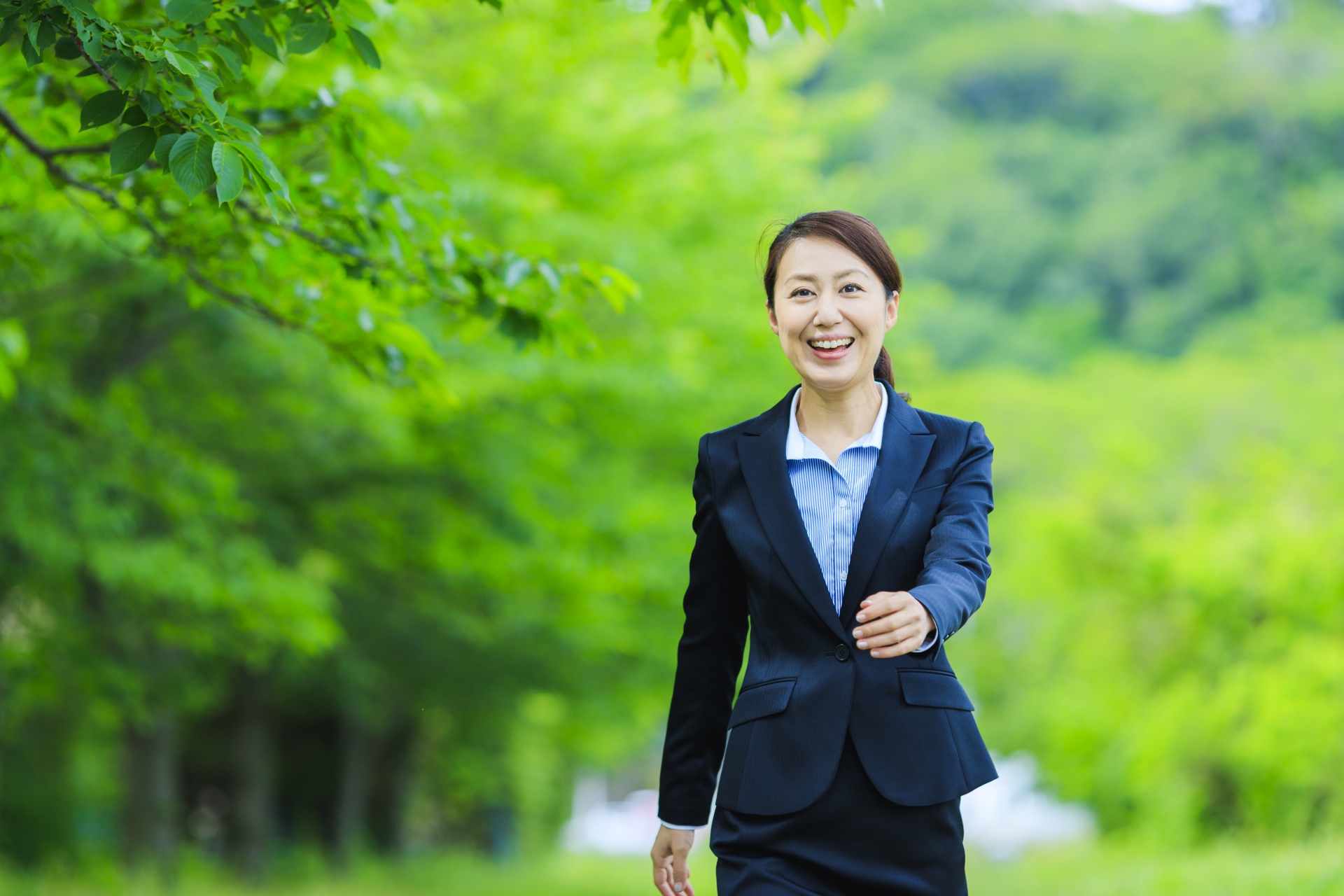 緑豊かな背景のなか、笑顔で前を向いて歩くリクルートスーツ姿の女性